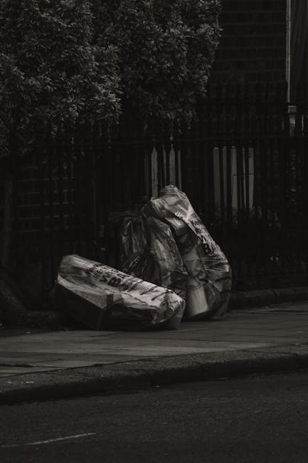 Several large black bin bags made of shiny, slightly crinkled plastic are placed on a paved sidewalk next to a black metal fence, which is partially obscured by dark foliage from a nearby tree. The bags are loosely stacked, with some leaning against each other, and appear to contain household waste or rubbish awaiting collection. The scene is set outdoors in a residential or urban area, with the background showing a dense cluster of trees or bushes and a dark, indistinct structure behind the fence. The lighting is low, giving a muted, monochromatic tone to the image. This visual depiction relates to waste management and private rubbish removal services, such as those provided by Waste Removal Streatham, highlighting the type of refuse handled during on-site clearance or alternative waste disposal processes common in non-public collection scenarios.
