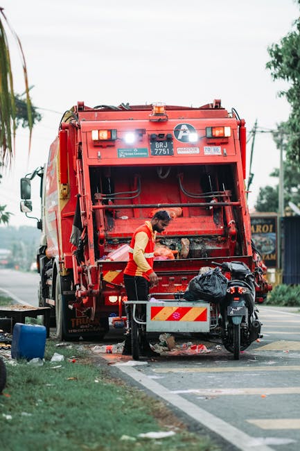 A sanitation worker dressed in a yellow and red uniform is standing next to a large red rubbish collection vehicle on a paved urban street, actively engaged in waste collection or cleaning activities. The back of the vehicle is open, revealing mechanical components and an empty rubbish compartment. The worker appears to be handling a black plastic bag, which is placed on a small metal platform attached to the vehicle. The scene includes a motorcycle parked nearby, partially obscured, with a black body and visible seat. In the background, there are trees, a utility pole with wires, and a blue building or shopfront with signage, indicating a commercial or residential area. The overall atmosphere suggests on-site waste management or rubbish removal, consistent with private or alternative waste handling services in Streatham, with natural daylight illuminating the scene and some green grassy patches adjacent to the street, emphasizing an outdoor environment focused on rubbish collection activities.